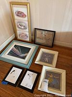 Photo of seven framed artworks of varied sizes and styles, arranged on hardwood floor against a wall.