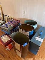 Boxes of glass Christmas ornaments with visible blue and white ornaments, two metal trash cans, and blue metal file box arranged on wood floor near wall.