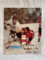 Front view of vintage color hockey photograph featuring Bobby Orr in white Boston Bruins uniform playing against Chicago Blackhawks players, with visible Nynex logo and black ink autograph.