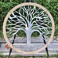 Full front view of large Tree of Life outdoor sculpture with silver galvanized steel tree in an unpainted round wooden frame, standing outside on a garden bench with greenery in the background.