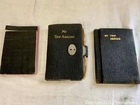Photo of three black travel diaries laid flat, two titled 'My Trip Abroad' with gold embossing, one plain smaller notebook.