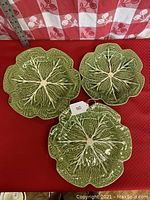 Set of three green cabbage leaf shaped ceramic plates on a red surface with a white and red checkered background and ruler for scale.