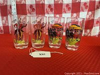 Four clear glass tumblers with vintage applied color graphics of bicyclist and horse-drawn carriage, photographed on red tablecloth with checkered background and measuring ruler for scale.