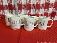 Front view showing 6 assorted Rae Dunn white ceramic mugs on red checkered tablecloth, each with unique lettering in black.