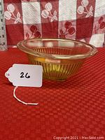 Side view of a small amber ribbed/fluted depression glass mixing bowl, showing the ribbed pattern, flared rim, and translucent amber color against a red patterned background.