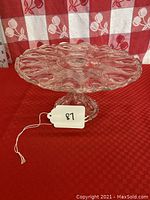 Side view of clear glass cake stand showing Peacock Feather pattern and full pedestal base on red surface with checkered background.