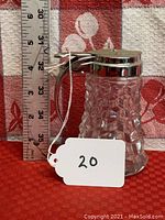 Side view of syrup pitcher with clear textured glass body and metal lid with handle. Ruler next to pitcher shows approximately 4 inches tall.