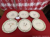 Six Lenox china plates arranged on red fabric background featuring Days 7 to 12 of the 12 Days of Christmas series, each depicting a holiday-themed scene with holly berry border.