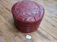 Top and angled side views of a single round burgundy mid-century Naugahyde ottoman with floral rosette stitching on top.