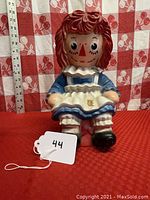 Front and side views of the Raggedy Ann coin bank sitting on a red background with a checkered cloth behind and a measuring tape beside showing approximate height of 9 inches.