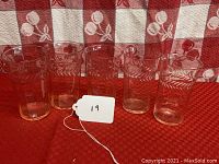 Set of 5 pink depression glass tumblers arranged in a row against red and white fabric, showing the translucent pink tint and etched laurel leaf band pattern.