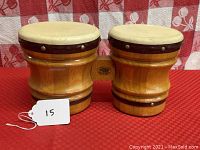 Pair of wooden bongo drums on red and white checkered surface. Both drums connected by a wooden bridge with visible marking, natural tan colored drum heads secured by metal tacks, light wood body with dark accent bands.