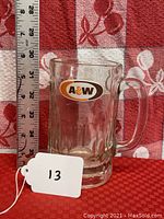 Two separate images showing clear glass mugs with the orange and brown A&W oval logo on the front. A measuring ruler confirms each mug is about 5.5 inches in height. The mugs have thick handles and ribbed bases.