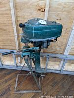 Side view showing green metal body with paint chipping and rust on the outboard motor mounted on a metal stand.