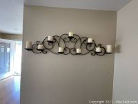 Wall mounted wrought iron candelabra with eight cream-colored pillar candles in place, mounted on beige wall.