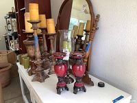 Photo of various wooden and metal candlesticks with wax candles, green glass votives, a large glass hurricane holder, and two red decorative candle holders on a white table in front of a mirror.