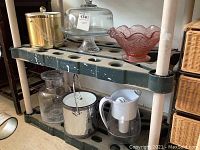 Photo showing the collection of glassware and ice buckets on a black shelf, including decorated pink bowl, cake stand, carafe, ice buckets, and filtered pitcher.