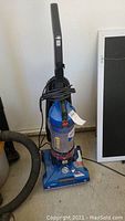 Front view of blue Hoover upright vacuum cleaner with power cord wrapped around handle and attachment hose.