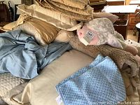 Overview of assorted pillows, blankets, and linens stacked on a surface showing beige and gold pillows, blue quilted linen, and multiple blankets.