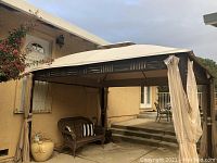 Outdoor metal patio gazebo assembled and shown on concrete patio next to house. Beige fabric cover with metal frame and tied side curtains seen.