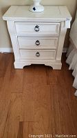 White painted wood nightstand with three drawers and black metal ring pull handles on a hardwood floor. The lamp on top is not included.