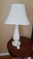 Full view of one white table lamp on wooden table showing square fabric shade and decorative embossed base.