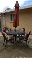 Full view of round metal table with umbrella center and four chairs with floral cushions on concrete patio against stucco wall