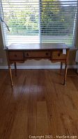Full view of the vintage wooden desk showing three drawers, turned legs, and scalloped apron in front of window.