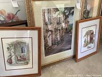Three framed wall art prints arranged together on the floor, featuring Mediterranean styled scenes including stone buildings, windows, and plants.