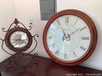 Wide photo of both clocks showing size and style. Wall clock is larger with wooden frame; mantle clock smaller with metal scroll frame.