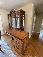 Wood desk with two drawers and metal handles, showing side profile and cable hole on surface. Wooden side cabinet positioned nearby.