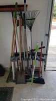 Front view of multiple yard tools held upright in a gray plastic shelving unit, including rakes, shovels, pruners, and a broom.