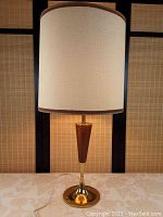 Full view of lamp showing tall cream-colored fabric shade and tapered teak and brass base.