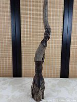 Full view of the tall slender carved hardwood sculpture showing the two carved human faces, one on the upper part and one on the lower part, with natural wood above and below each face.