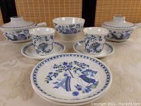 Full view of 11 piece ceramic dinnerware set with stylized blue and white handpainted floral and figure design, showing 2 covered bowls with handles, teacups, saucers, small plates and bowl.
