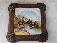 Close-up of antique brass trivet with hand-painted tile showing Invegarry Castle and lake view with a boat.