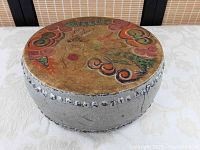 Front view of round antique wooden drum with handpainted colorful leather drumhead and metal tacks securing the skin around the rim.