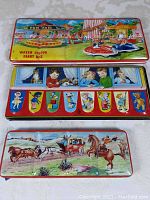 Top view of three closed vintage watercolor paint tins with lithograph illustrations: fun fair bumper cars scene, children and cartoon flags, and stagecoach western scene.