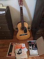 Full view of the Westbrook acoustic guitar standing upright against a wall with accompanying music books and VHS tape displayed nearby on the floor.