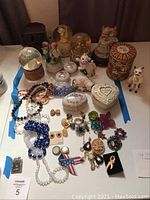 Full lot showing snow globes, cat figurines, various costume jewelry, and ceramic jewelry holders arranged on a surface.
