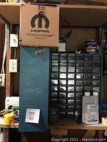 Overview of garage shelf showing two hardware organizers, black with many small drawers and blue organizer with fewer drawers. Surrounding miscellaneous garage items and a cardboard Mopar box on top.