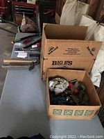 Overview of tools and boxes on a table in a garage setting including hammers, branch nippers, and boxes with miscellaneous items and tie down straps.