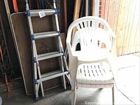 Photo showing aluminum extension ladder folded for storage leaning against wall with folding camp table and stacked white plastic chairs nearby.