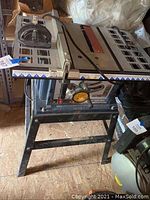 Photo showing the full Ryobi 10 inch table saw mounted on black metal stand in a shed environment, with measurement markings on the table surface and power cable visible.