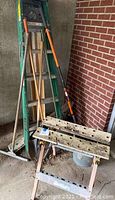 Adjustable sawhorse in worn and dirty condition alongside green folding ladder and several yard tools with wooden handles leaning against a brick wall.
