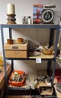 A shelving unit holding assorted vintage kitchen items including glassware, silverplate serving pieces, ceramic hen containers, a box of flatware, and various smaller kitchen tools and accessories.