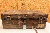 Front angled view of antique steamer trunk showing leather covering, metal reinforcements, travel stickers, and central lock.