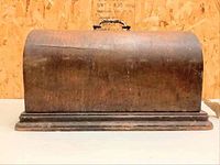 Closed view of the antique sewing machine wooden case from front side, showing curved top and handle.