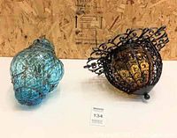 Two glass pendant lights, one blue with metal wire frame and one amber with black metal scrollwork frame, placed on a surface with a wooden wall background.