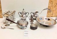 Various silver and silverplated servingware including teapot, pitcher, trays, stemmed glasses, pans, and spoons arranged on table.